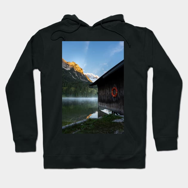 Boathouse with lifesaver Portrait. Amazing shot of a wooden house in the Ferchensee lake in Bavaria, Germany, in front of a mountain belonging to the Alps. Scenic foggy morning scenery at sunrise. Hoodie by EviRadauscher